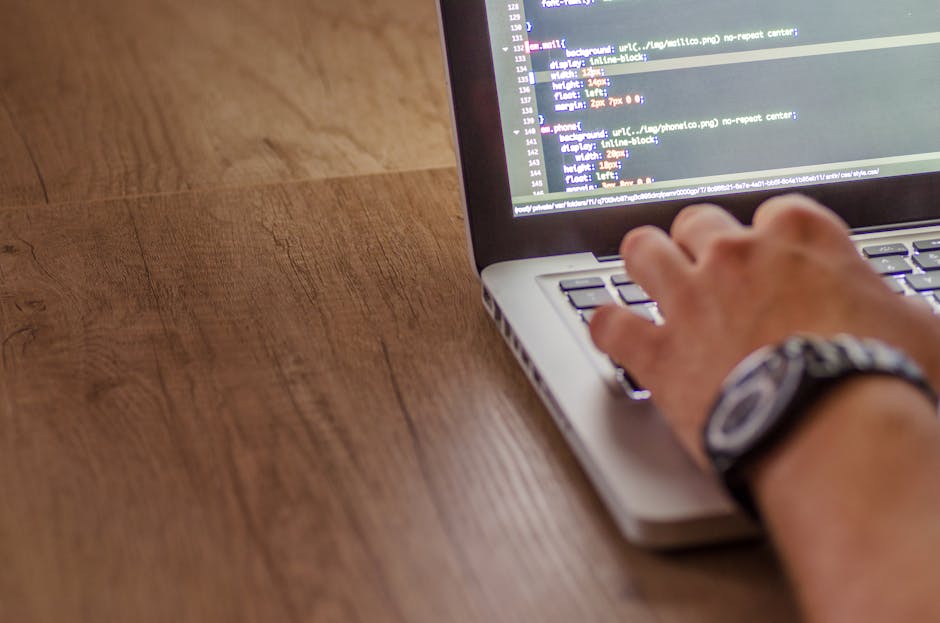 Close-up of a person's hand operating a laptop on a wooden desk, displaying lines of programming code or website development scripting. The individual is wearing a watch with a dark band on their wris