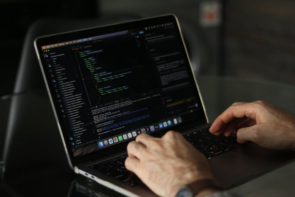 Close-up of a person's hand operating a laptop on a wooden desk, displaying lines of programming code or website development scripting. The individual is wearing a watch with a dark band on their wris